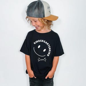 Könnte beinhalten: Ein schwarzes T-Shirt mit einem weißen Grafikdruck eines lächelnden Gesichts, dem Text "KINDERGARTENKIND" in einem Bogen darüber und dem Namen "Leon" darunter, kombiniert mit einer grau-beigen Trucker-Cap mit Netz und beigem Schirm.