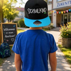 Könnte beinhalten: Eine schwarze Snapback-Cap mit einem hellblauen Schirm. Auf der Vorderseite der Kappe steht der weiße Schriftzug SCHULKIND und der grüne Text #2026. Ein kleiner schwarzer Aufkleber mit dem Wort Snapback ist mittig auf dem blauen Schirm platziert.