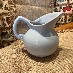 May include: A light blue ceramic pitcher with a rounded body, a wide pouring spout, and a curved handle. The pitcher features a smooth, glossy finish and a solid, uniform color throughout its exterior surface, showcasing a simple, functional design.