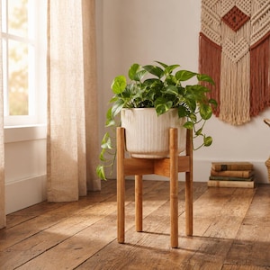 May include: A white ceramic planter with vertical ribbed detailing sits inside a light brown wooden plant stand. The stand features four cylindrical legs connected by a cross-shaped support. A green leafy plant grows from the planter.