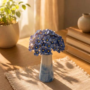 Peut inclure: Un vase en céramique avec une finition dégradée bleue et blanche, contenant un bouquet de petites fleurs bleues artisanales aux centres jaune vif. Les fleurs sont disposées en un bouquet dense et arrondi, créant un arrangement floral éclatant.
