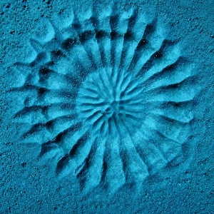 May include: A vibrant blue textured surface featuring a deeply embossed circular pattern. The design consists of radiating ridges that extend from a central point, creating a sunburst or floral-like impression with distinct shadows and highlights.