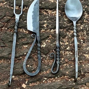 May include: A set of four hand-forged metal utensils including a two-pronged fork, a curved blade knife with a looped handle, a thin pointed tool, and a spoon. Each piece features a twisted metal handle design with a dark, textured finish.