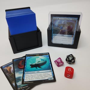 May include: A black plastic card deck box containing a stack of blue-sleeved cards, a second black holder displaying a Japanese trading card in a clear sleeve, three loose trading cards, a magenta twenty-sided die, and a red six-sided die.