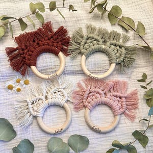 Puede incluir: Cuatro anillos circulares de madera con cordón de macramé en color óxido, verde salvia, crema y rosa empolvado. Cada anillo tiene un diseño de flecos anudados y está personalizado con los nombres Leonie, Hannes, Julian y Emily grabados.
