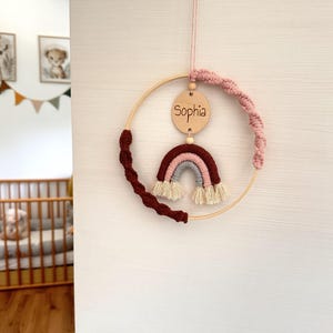 Puede incluir: Un adorno de pared circular de madera con un arcoíris de macramé en tonos rojo oscuro, rosa y gris con flecos crema. Un disco de madera grabado con el nombre Sophia cuelga de la parte superior, acentuado por cordones trenzados rosa y rojo.