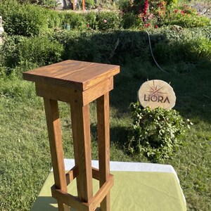 May include: A tall, square-topped wooden pedestal stand with a dark brown stained finish. The stand features four vertical legs connected by a lower support frame, showcasing a visible wood grain texture throughout the entire structure.