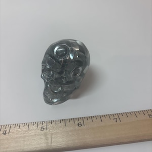 May include: A translucent dark grey skull figurine with a faceted geometric surface texture. The skull is positioned next to a wooden ruler, showing it measures approximately 3.8 cm in width at its widest point near the top.