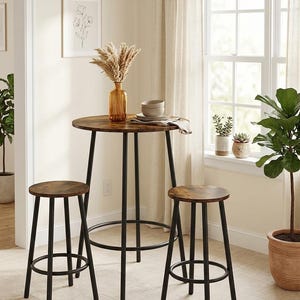 May include: A tall round bistro table with a dark wood grain top and black metal legs, accompanied by two matching round stools with black metal frames. The table holds a glass vase with dried pampas grass and a stack of two ceramic bowls.