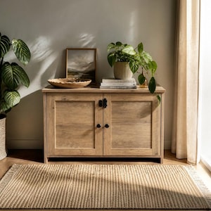 May include: A light brown wooden cabinet with two doors, black knobs, and black hinges. On top sits a shallow wooden bowl, a framed landscape print, and a potted green plant resting on a stack of two books with white spines and black text.