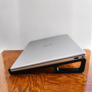 Puede incluir: Un portátil plateado con el logotipo de HP en la tapa está colocado en ángulo sobre un soporte negro de marco abierto. El portátil cuenta con un puerto USB y una toma de auriculares visibles en el lateral, apoyado sobre la estructura.
