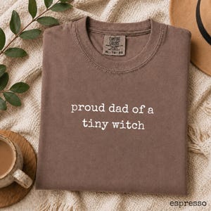 May include: A brown short-sleeved t-shirt with a crew neckline and the white text "proud dad of a tiny witch" printed on the chest. The shirt features a Comfort Colors brand label on the inside collar with the size XL-TG-EG clearly visible.
