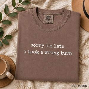 May include: A brown short-sleeve t-shirt with a crew neckline and the white text "sorry i'm late i took a wrong turn" printed on the front. The shirt features a Comfort Colors brand label on the inside collar with the size marked as XL.