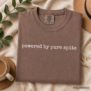以下が含まれることがあります： 胸元に白い文字で「powered by pure spite」とプリントされた茶色の半袖Tシャツ。シャツの内側のネックラインにはComfort Colorsのブランドラベルがあり、ラベルにはサイズXL、TG、EGが記載されています。生地は無地です。