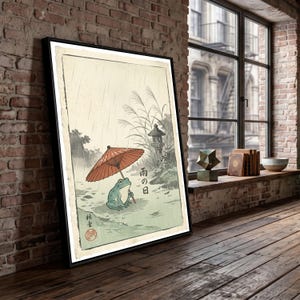 以下が含まれることがあります： 雨の中で赤い和傘をさした緑色のカエルの絵が描かれた額入りアートプリント。イラストには背の高い草、石灯籠、そして「雨の日」と書かれた日本語の文字が含まれており、全体が落ち着いた質感の背景に配置されています。