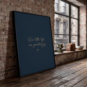 May include: A framed vertical wall art piece featuring a dark navy blue background with the gold-colored cursive text "Our little life, our greatest joy." and a small heart symbol centered below the text, enclosed in a thin black frame.