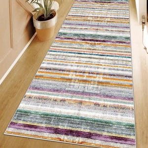 May include: A long rectangular runner rug featuring a multi-colored horizontal striped pattern. The design incorporates bands of purple, orange, teal, grey, and white with a distressed, weathered texture throughout the entire surface of the rug.