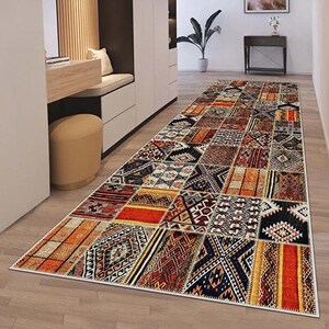 May include: A long rectangular runner rug featuring a patchwork design of various geometric and tribal patterns in shades of orange, red, black, white, and blue, laid out on a light wood floor in a hallway with white walls and modern furniture.