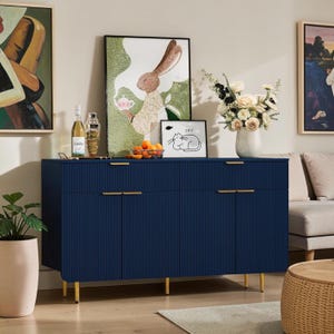 May include: A navy blue sideboard with a vertical ribbed texture, four doors, and slim gold handles. The piece stands on thin gold legs and features a flat top surface holding a glass bottle, a bowl of oranges, a framed rabbit print, and a vase of flowers.