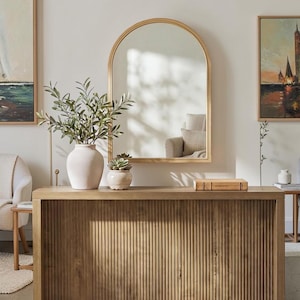 May include: A light wood console table with a vertical fluted front design, topped with a white vase containing green branches, a small succulent, and a book. Above the table hangs an arched mirror with a thin gold frame on a plain wall.