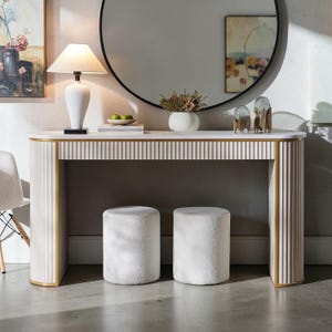 May include: A white console table with a fluted front and gold trim, featuring a smooth rectangular top. Two matching white cylindrical stools with textured fabric are positioned underneath the table, which is supported by curved, fluted side panels.