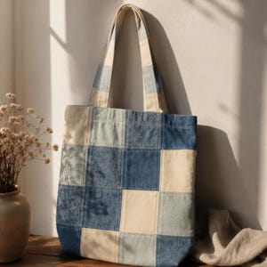 Può includere: Una borsa tote realizzata con un patchwork di pannelli di tessuto quadrati in varie tonalità di blu, denim e crema. La borsa presenta due lunghi spallacci e un corpo rettangolare con un visibile motivo a griglia di quadrati di tessuto cuciti.