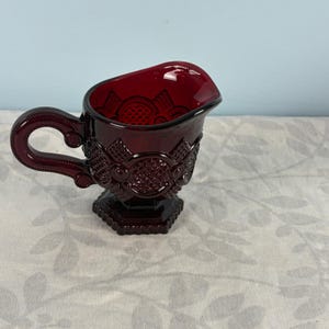 May include: A deep red glass creamer jug featuring a pressed diamond and circle pattern across the exterior. The vessel has a curved handle, a wide pouring spout, and a hexagonal base, showcasing a translucent, rich ruby-coloured finish.