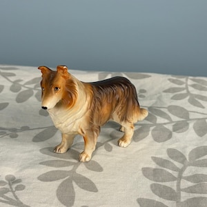May include: A ceramic figurine of a collie dog standing in a profile position. The dog features a coat with shades of golden brown, tan, and white, with dark brown accents along the back and ears. The figurine has a smooth, glossy finish.