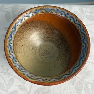 May include: A round ceramic bowl with a speckled beige interior that transitions to a warm orange-brown rim. The outer edge features a decorative blue and white wavy pattern embossed into the clay, creating a textured border.