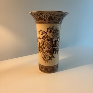 May include: A cream-colored ceramic vase with a flared rim, featuring a dark brown floral pattern around the top and bottom borders, and a central bouquet of detailed flowers and leaves in the same dark brown tone on the main body.
