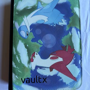 Pode incluir: Um fichário retangular para cartas colecionáveis com lombada preta e capa frontal exibindo um Latios azul e branco e uma Latias vermelha e branca voando sobre um mapa terrestre estilizado em verde e azul. O texto vaultx está impresso em preto na parte inferior.