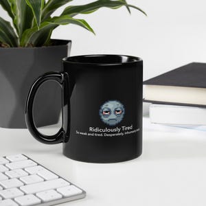 May include: A black ceramic mug featuring a pixel art graphic of a tired, grey, round face with red-rimmed eyes. Below the graphic, the text reads: Ridiculously Tired. So weak and tired. Desperately, inhumanly tired. The mug has a curved handle.