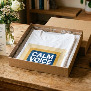 May include: A white t-shirt folded inside a brown cardboard box. The shirt features a graphic design with a mustard yellow rectangular border and a dark blue centre containing the white text "CALM VOICE" in a bold, sans-serif typeface.