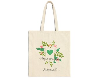 Op de afbeelding: Een beige canvas draagtas met een afbeelding van een wit t-shirt op een hanger met een groen hart, omringd door groene bladeren en roze bloemen. De tekst "Hope springs" is gedrukt boven de tekst "Eternal....." in een donker cursief lettertype.