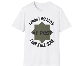 Op de afbeelding: Een wit t-shirt met korte mouwen en een olijfgroene achtpuntige stergrafiek in het midden. De tekst "I KNOW I AM LOVED" staat in een boog boven de ster, "#1 PEST" staat in het midden van de ster en "I AM STILL ALIVE" staat in een boog eronder.
