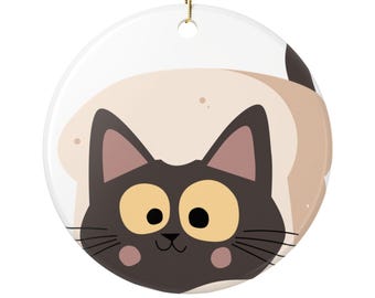 Op de afbeelding: Een rond wit keramieken ornament met een cartoonillustratie van een donkerbruine kat met grote gele ogen en roze wangen, geplaatst voor een snee beige brood, compleet met een gouden ophangkoord aan de bovenzijde.