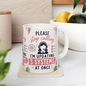 May include: A white ceramic mug featuring a graphic of a person wearing a headset with the text "PLEASE stop calling, I'M UPDATING 3 SYSTEMS AT ONCE". The design includes pink and dark blue lettering, a speech bubble with a scribble, and checkboxes.