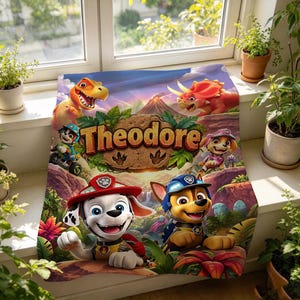 May include: A rectangular fleece blanket featuring a colorful cartoon scene with Paw Patrol characters, a T-Rex, and a Triceratops. The name Theodore is printed in large, textured brown letters in the center above a volcanic landscape.