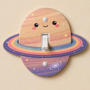 Puede incluir: Una placa de interruptor ovalada con un planeta de dibujos animados que tiene una cara sonriente, mejillas rosadas y anillos coloridos. El diseño incluye pequeñas estrellas amarillas y una galaxia espiral. Un interruptor blanco muestra el texto OFF.