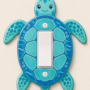 Peut inclure: Une plaque d'interrupteur en forme de tortue de mer dans des tons bleu et sarcelle. La tortue présente un visage souriant, des nageoires à motifs et une carapace encadrant un interrupteur à bascule blanc fixé par deux vis blanches.
