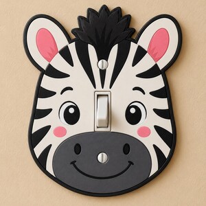 May include: A light switch plate shaped like a zebra head with black and white stripes, pink inner ears, and pink cheeks. The face features a dark grey muzzle with a smiling mouth and two small nostrils. A white toggle switch is centred in the middle.