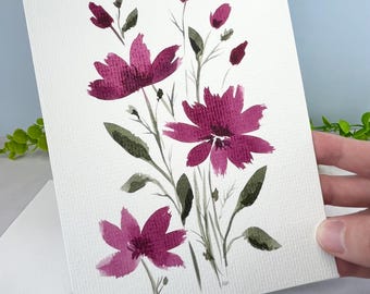 May include: A rectangular greeting card featuring a watercolor painting of magenta flowers with dark green leaves and stems on a textured white paper background. The floral illustration includes three open blossoms and several small buds.