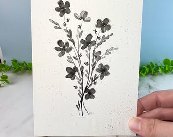 May include: A rectangular greeting card featuring a black ink watercolor illustration of a wildflower bouquet. The flowers and leaves are rendered in varying shades of gray and black on a textured off-white paper with subtle gray speckles.