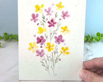 May include: A rectangular greeting card featuring a watercolor illustration of a wildflower bouquet. The artwork displays yellow and pink flowers with green stems and leaves on a white background with subtle gray speckles. The initials KD are visible.