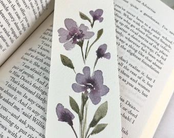 May include: A rectangular bookmark featuring a watercolor painting of purple flowers with green leaves on a white background. The floral illustration displays three main blossoms and several buds arranged vertically on thin stems with small foliage.