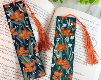 May include: Two rectangular bookmarks with a dark blue background featuring a painted pattern of orange flowers and green leaves. Each bookmark has a hole at the top with an attached orange tassel. The bookmarks are resting on an open book.