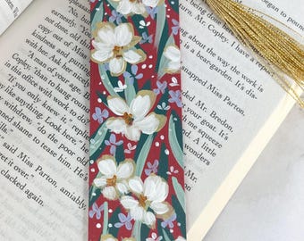 May include: A rectangular bookmark with a deep red background featuring hand-painted white flowers, green leaves, and small light purple accents. The bookmark includes a gold-colored metallic tassel attached to the top with a thin cord.