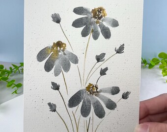 May include: A rectangular greeting card featuring a watercolor painting of three flowers with dark gray petals and golden yellow centers. The stems are thin and light brown, and the off-white paper background has subtle gray paint speckles.