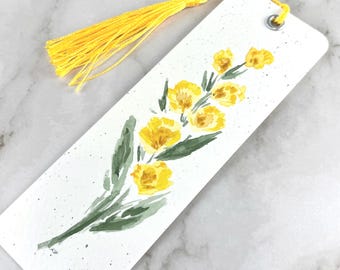 May include: A rectangular white bookmark featuring a watercolor painting of yellow flowers with green leaves. The bookmark has a silver metal eyelet at the top with a bright yellow tassel attached. The background of the bookmark has small black speckles.