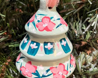 May include: A three-tiered, bell-shaped ornament in light mint green, hand-painted with coral pink flowers, dark teal rectangular accents, and small white floral details. A frayed coral pink ribbon is attached to the top for hanging.